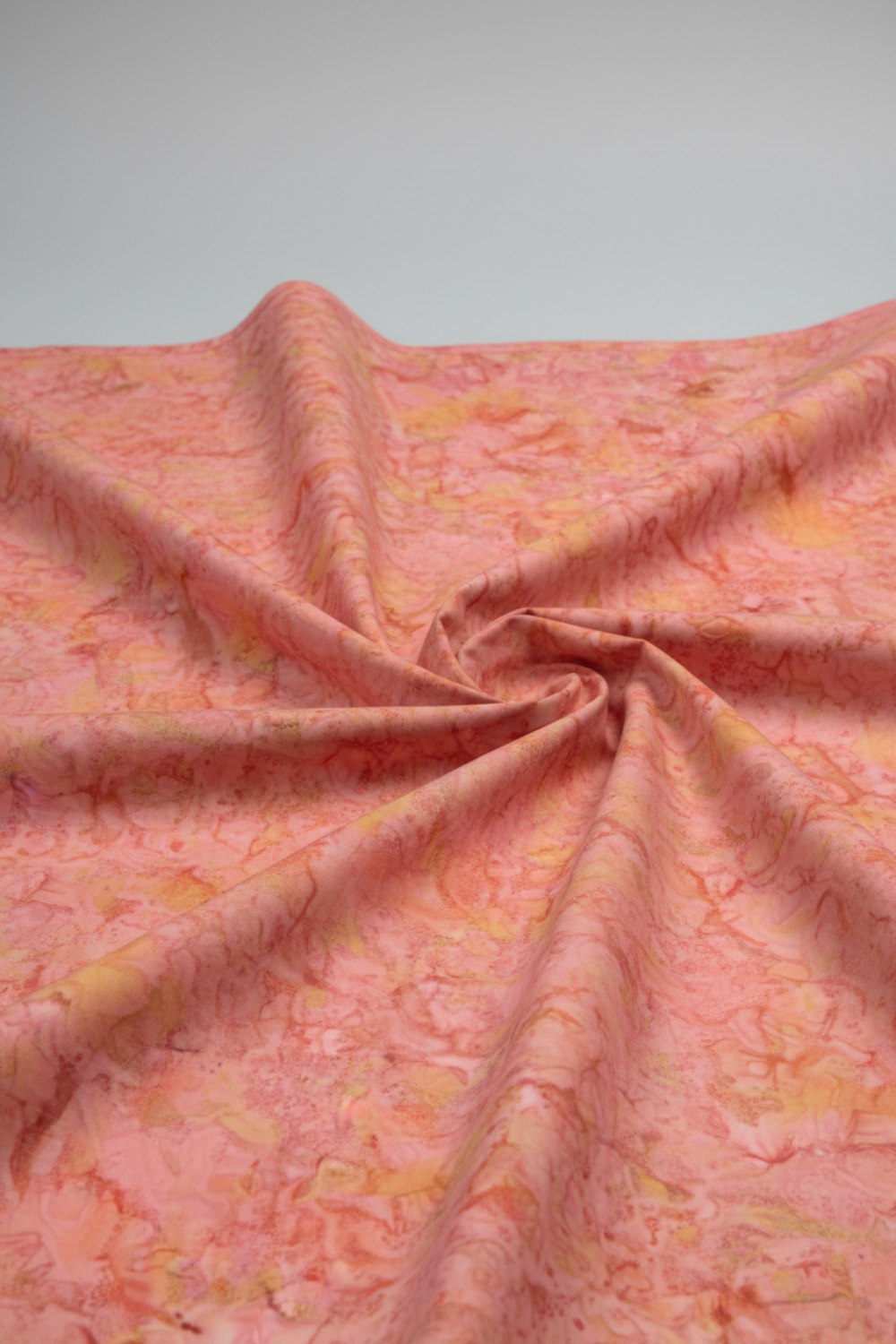 Expressions Batiks Hand-Dyed Sunset quilting cotton fabric by Riley Blake Designs – warm coral pink, golden yellow, and peach hand-dyed batik in soft folds on light grey background emphasizing texture and drap