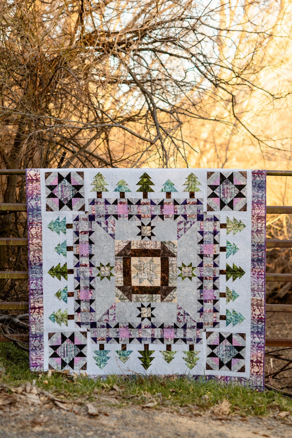 A Walk in the Woods quilt displayed flat against a rustic fence outdoors — purple, teal, green, pink, and cream Expressions Batiks by Riley Blake Designs — churn dash, star, and tree block design by Beverly McCullough — pattern available at IraRobi Quilts