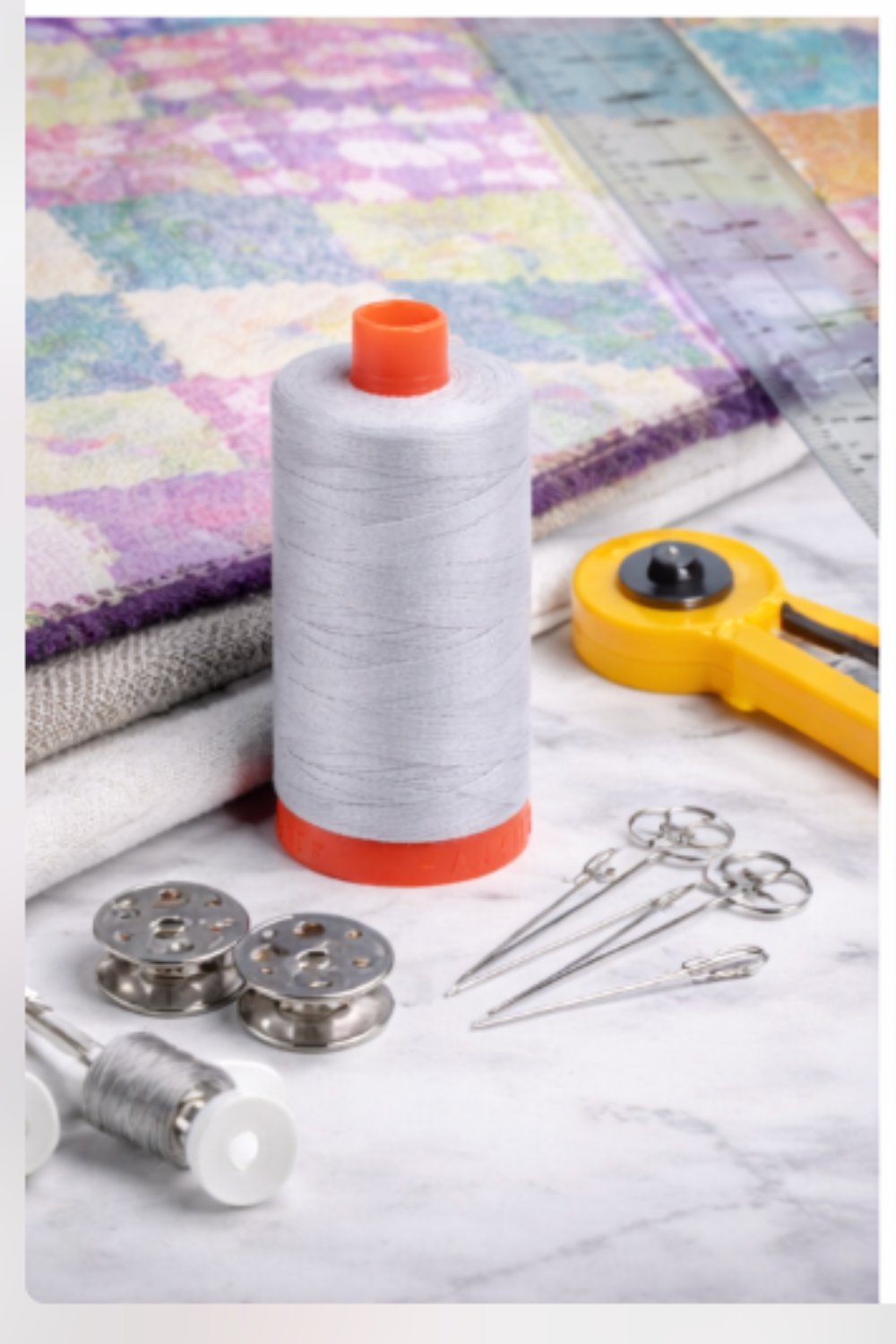 Aurifil Mako Cotton Thread 50wt Dove surrounded by rotary cutter, needles, and quilting tools.