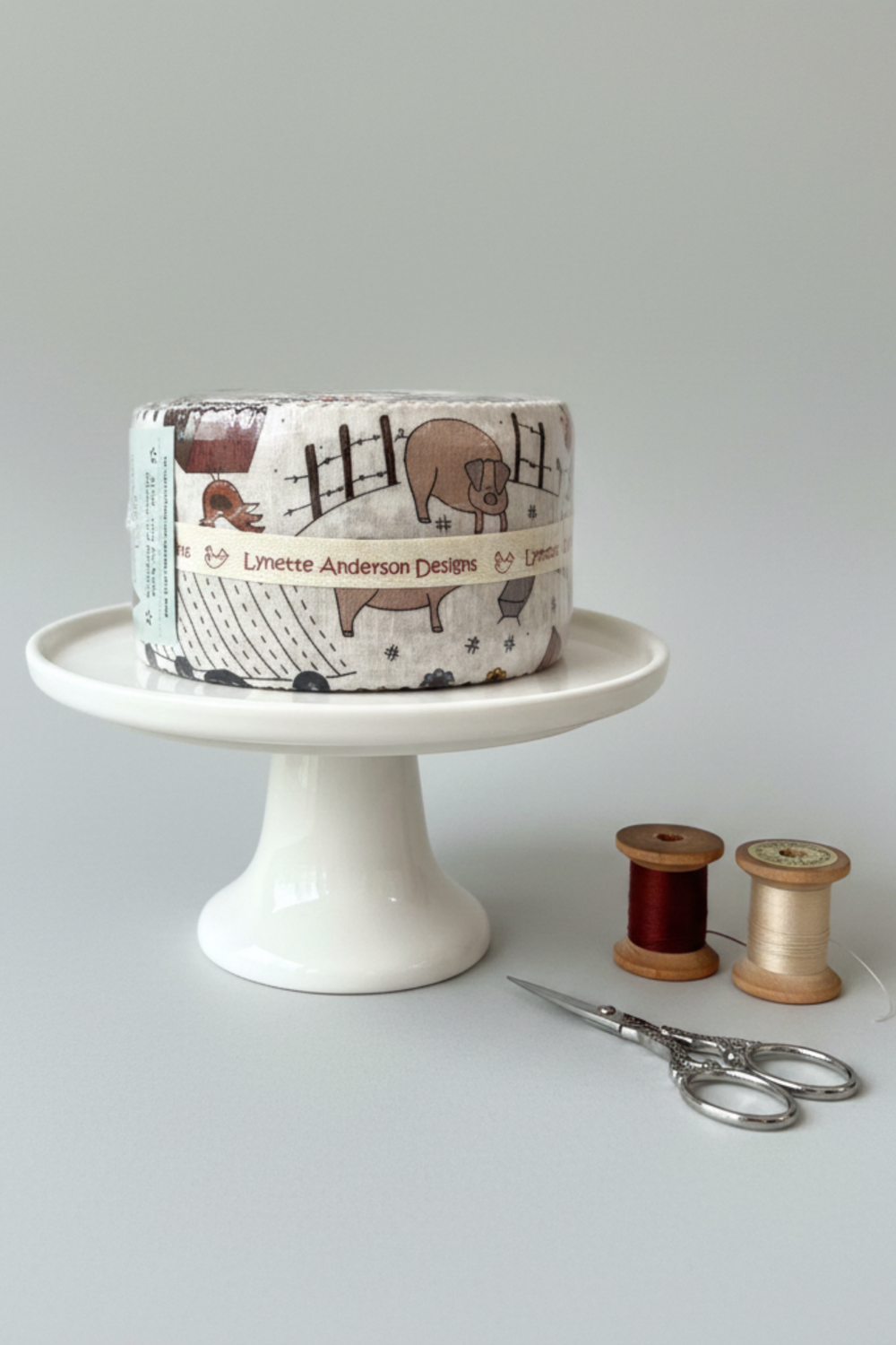 High Meadow Farm Jelly Roll by Lynette Anderson Designs displayed on white cake stand with appliqué scissors and wooden thread spools