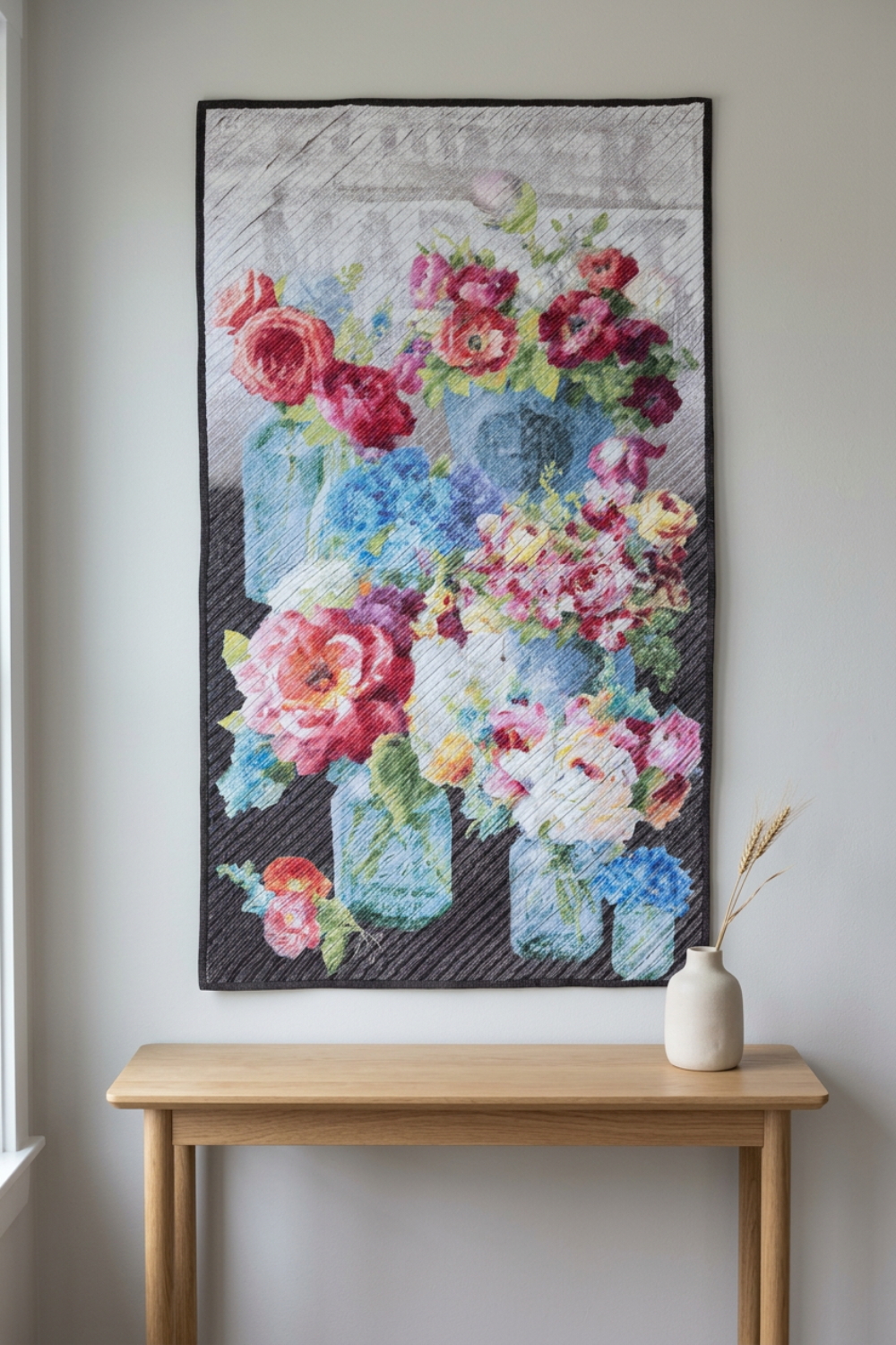 Vase with Flowers floral wall quilt hanging above an oak console beside a window, with a cream vase holding dried stems.