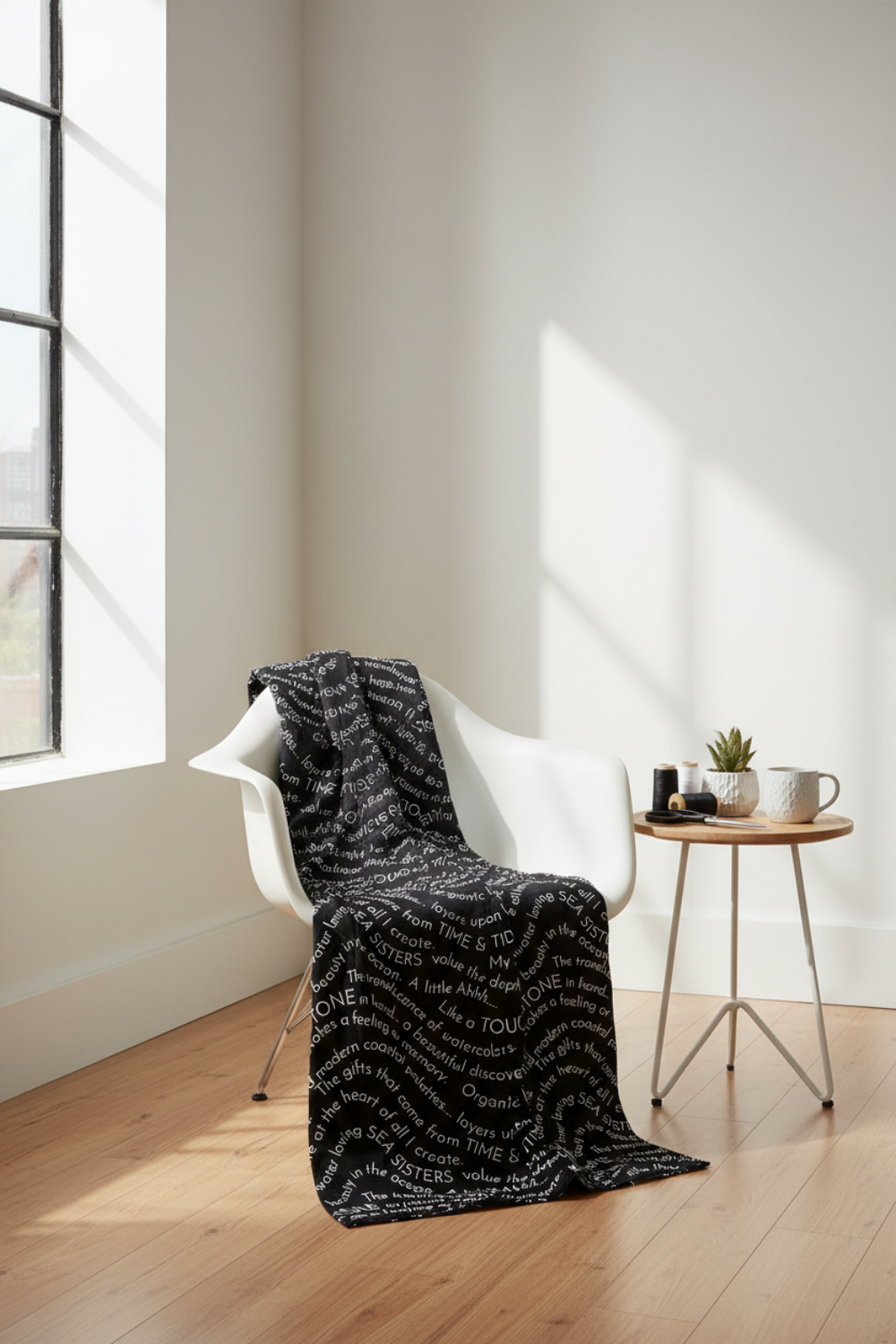 Writing on the Wall Script Black quilting cotton by FreeSpirit Fabrics -- bold black script fabric draped over a white modern chair in a bright loft sewing studio near a large window, with thread spools, scissors, and ceramic mug on a side table