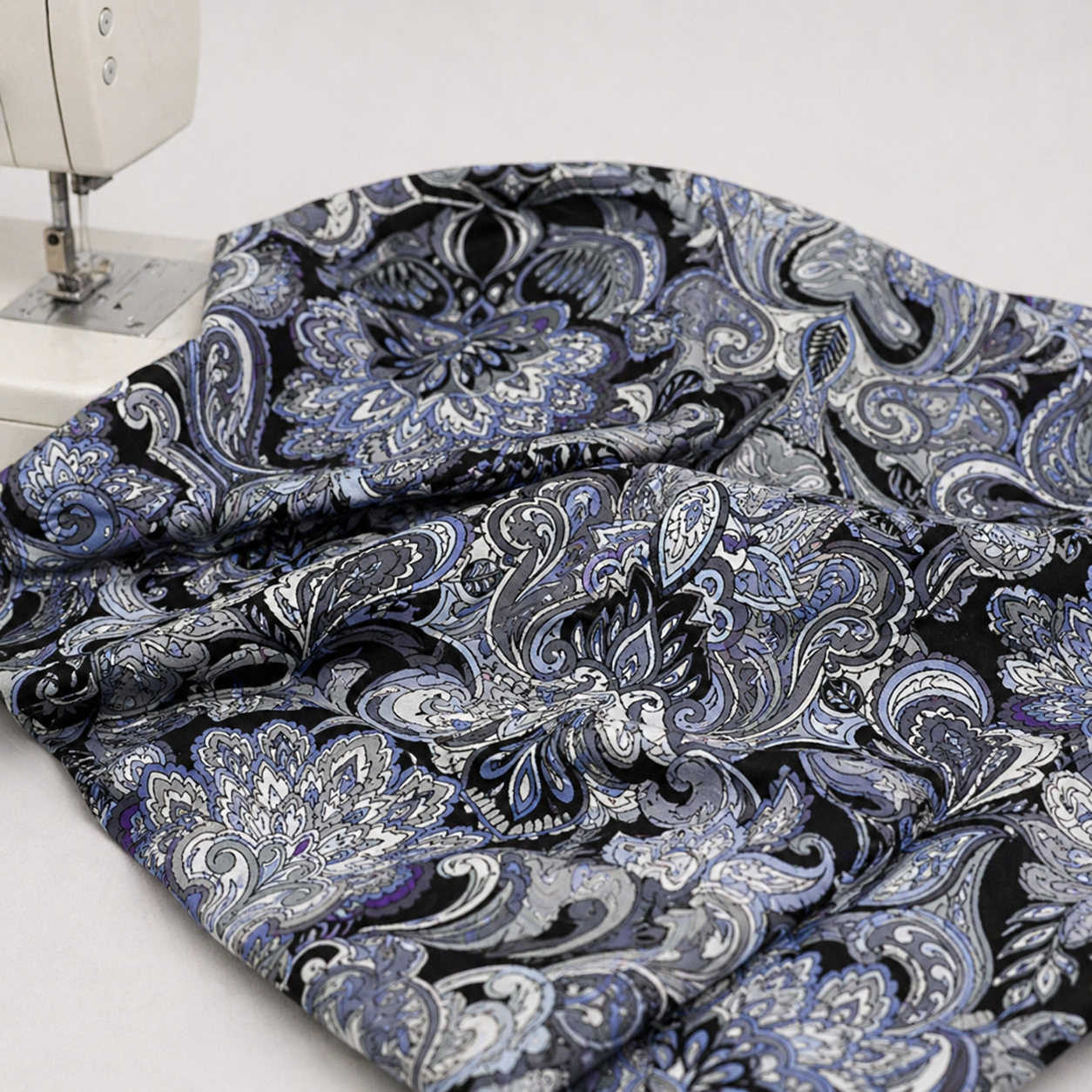 What If Prima Donna Grey/Blue baroque damask quilting fabric draped at sewing machine, showing paisley and floral medallion print in black, periwinkle blue, slate grey, and purple — Paula Nadelstern for Benartex