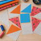 Safari Fun Floating Triangles White/Black by Terry Runyan for Benartex – cut half-square triangle quilt blocks in progress on a wooden table, featuring white with black triangle outlines paired with orange, blue, and coral parrot Safari Fun fabrics, with rotary cutter, pins, and thread spools