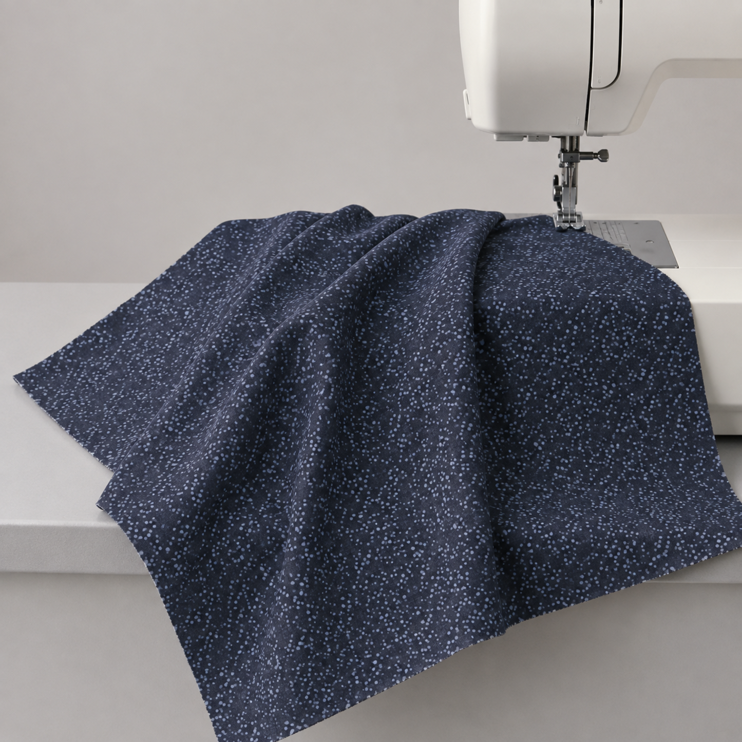 Dotty Thatched Indigo fabric by Robin Pickens for Moda Fabrics draped over a sewing machine with soft natural folds showing the deep indigo-charcoal ground with scattered tonal grey dots. Available at IraRobi Quilts.