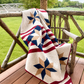 Fifty Nifty finished throw quilt by Lisa Lund for Shadow Valley Quilts — patriotic Lone Star blocks in navy and tan on diagonal red and cream striped sashing, draped over rustic twig chair on wooden porch with green meadow and bluebonnets in background — throw size 60″×72″