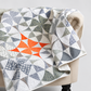 Chic Country finished quilt by Sew Kind of Wonderful — modern curved pinwheel blocks in scrappy gray, white, and low-volume prints with a single bold orange accent block, draped over a cream upholstered armchair — striking modern quilt design using the Quick Curve Ruler