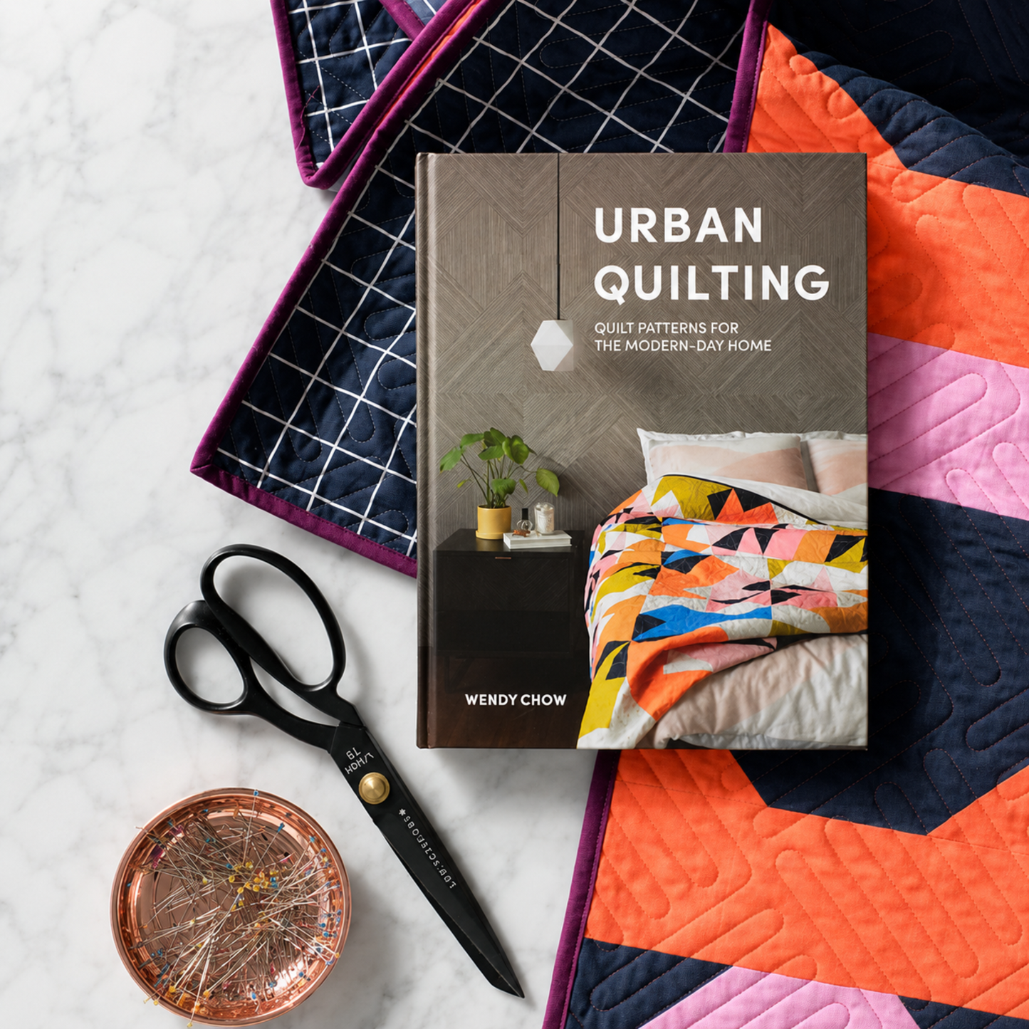 Urban Quilting Book by Wendy Chow hardcover on marble surface surrounded by black fabric scissors, copper pin dish, navy grid quilt, and bold orange and pink geometric quilt — Quilt Patterns for the Modern-Day Home