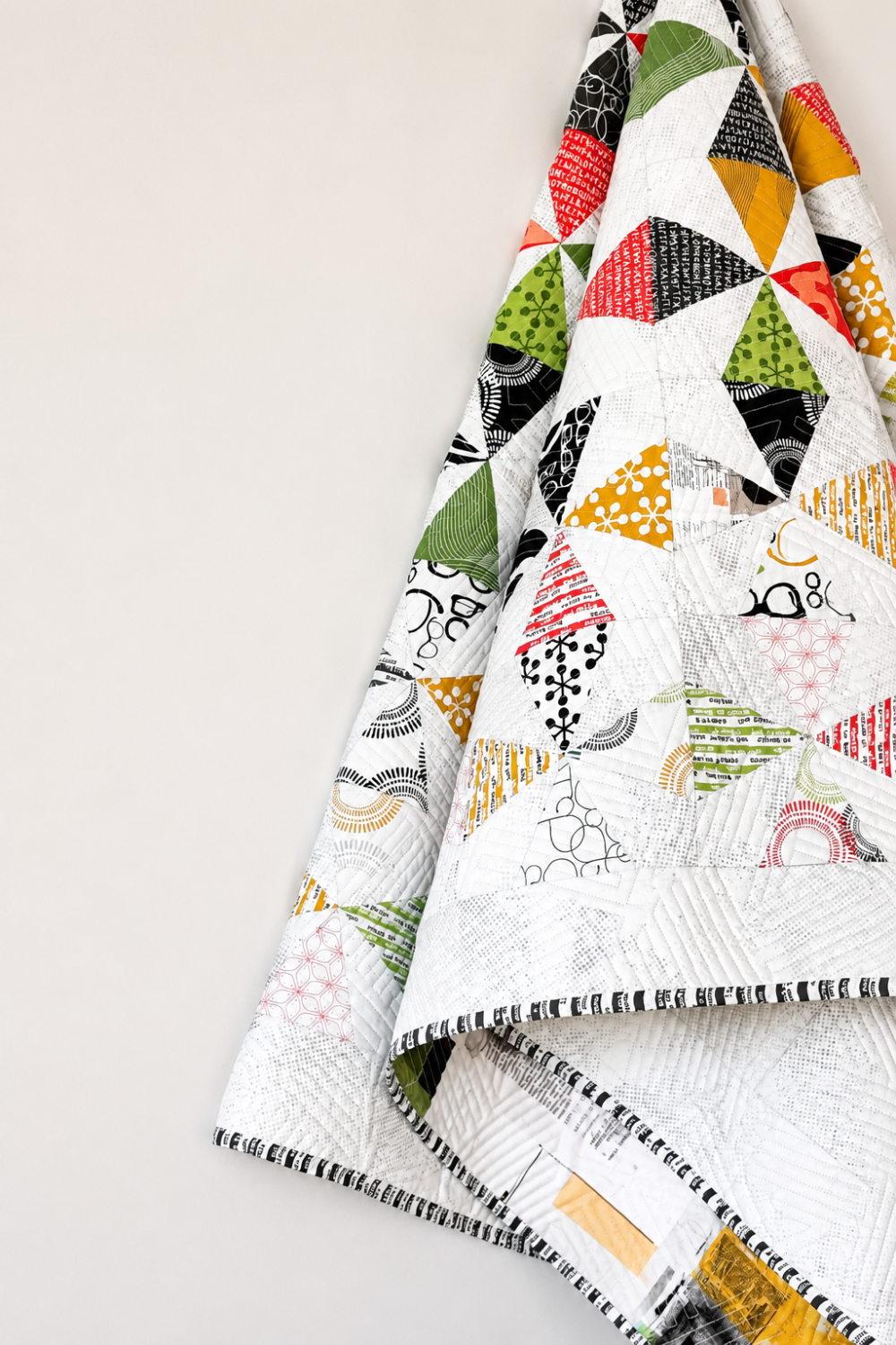 Dissolve quilt pattern by Zen Chic — close-up of FPP quilt hanging on wall showing bold geometric diamond and triangle fragments in red, green, gold, black and white Ampersand prints with whisper-white background and black-and-white striped binding — Dissolve pattern available at IraRobi Quilts