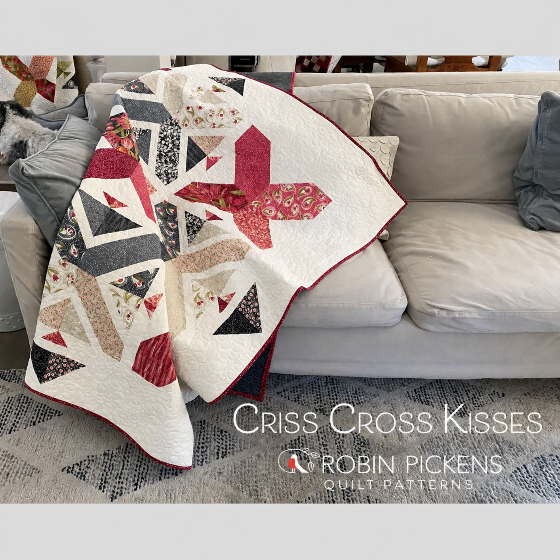 Criss Cross Kisses quilt by Robin Pickens draped over gray sofa showing X blocks and arrowhead details in crimson, navy and floral prints on cream background with red binding