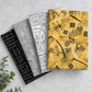 Writing on the Wall quilting cotton collection by FreeSpirit Fabrics -- three coordinating fabrics fanned on a marble surface with eucalyptus, showing black word collage, grey and white Well Worded word collage, and golden yellow abstract scratch and grid print -- modern graphic text prints, 100% cotton