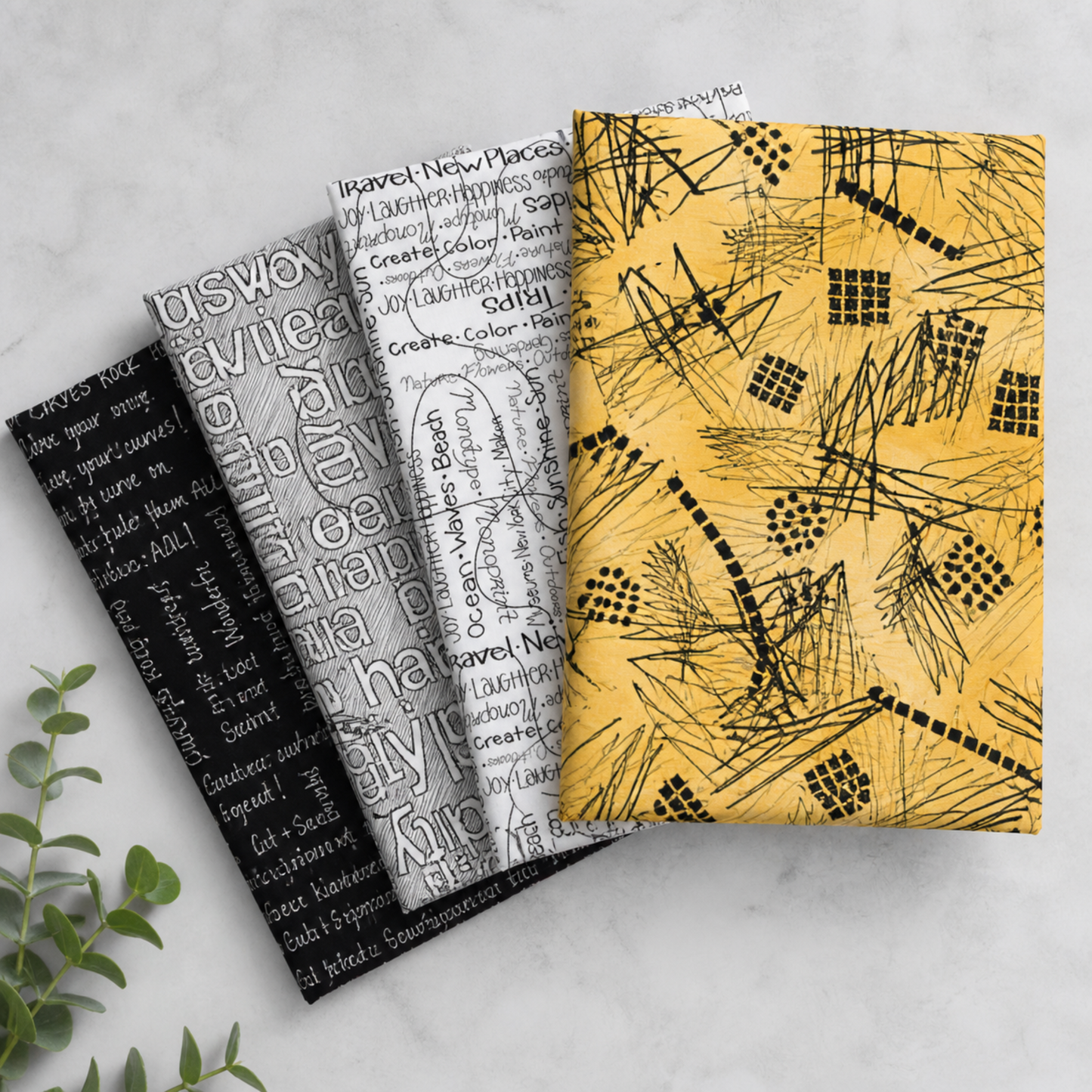 Writing on the Wall quilting cotton collection by FreeSpirit Fabrics -- three coordinating fabrics fanned on a marble surface with eucalyptus, showing black word collage, grey and white Well Worded word collage, and golden yellow abstract scratch and grid print -- modern graphic text prints, 100% cotton