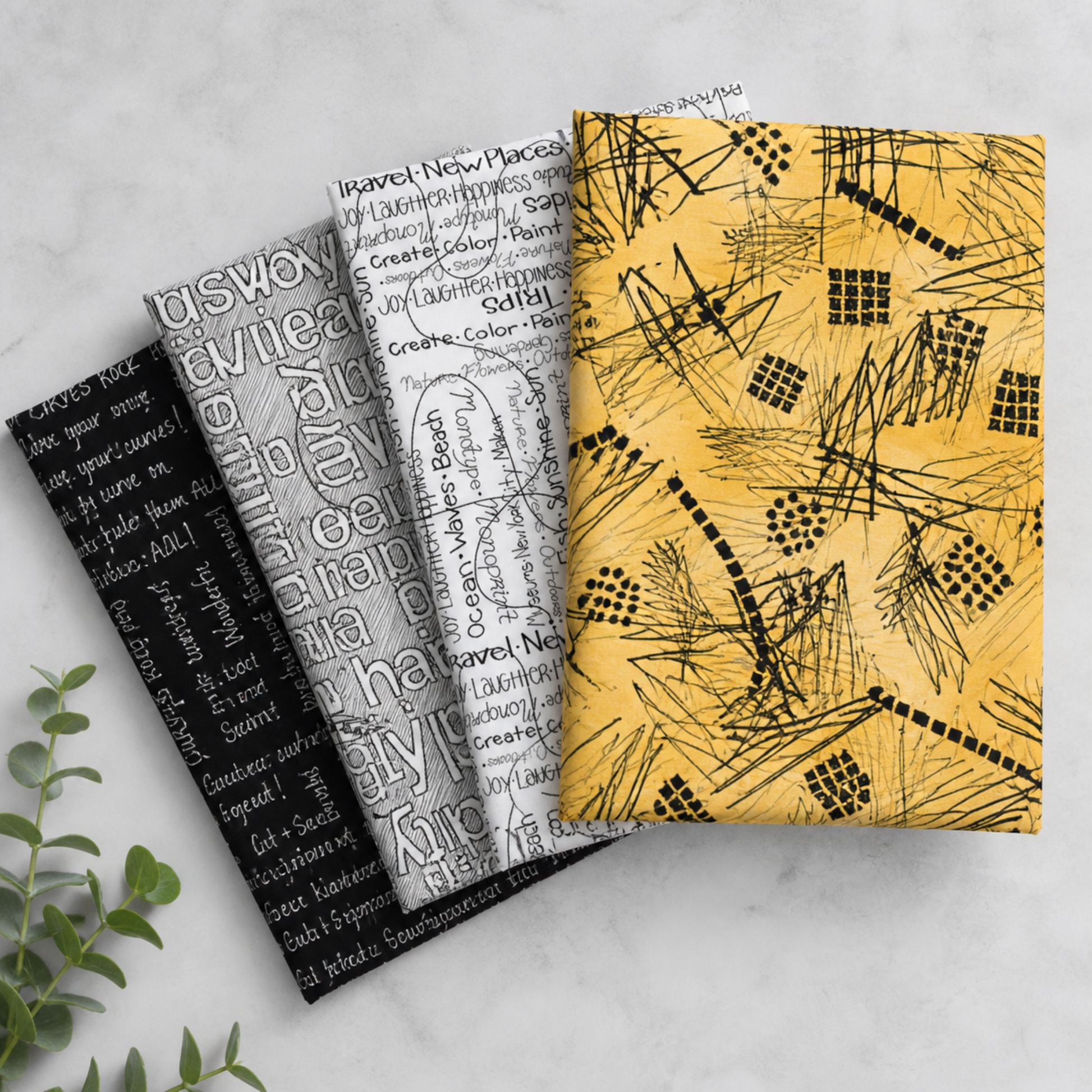 Writing on the Wall quilting cotton collection by FreeSpirit Fabrics -- three coordinating fabrics fanned on a marble surface with eucalyptus, showing black word collage, grey and white Well Worded word collage, and golden yellow abstract scratch and grid print -- modern graphic text prints, 100% cotton