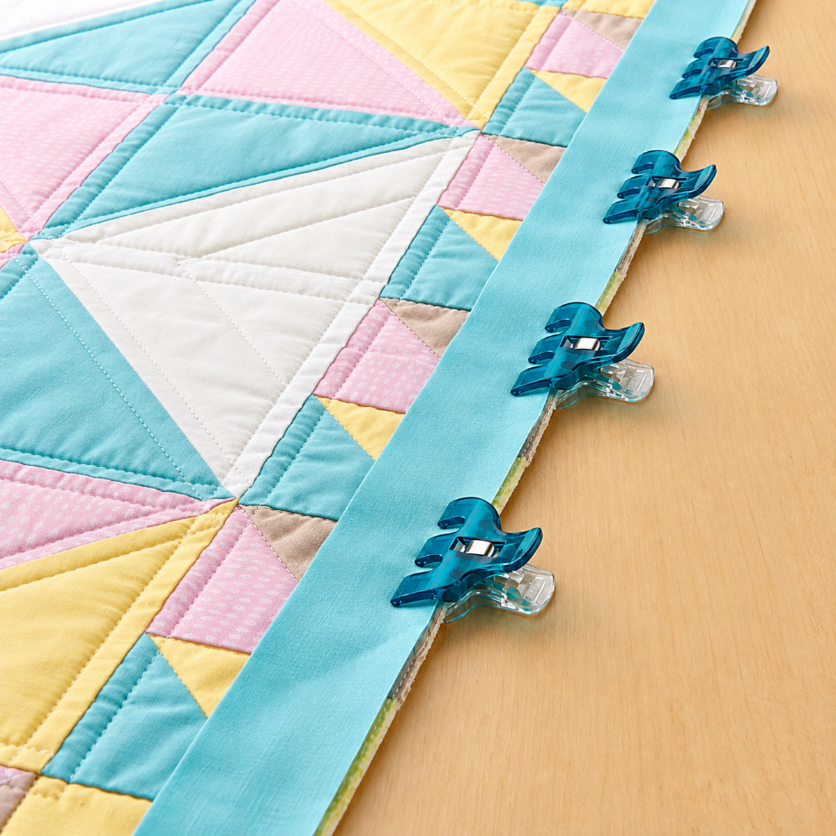 our Clover Triple Wide Wonder Clips securing a blue quilt binding strip to a pastel patchwork quilt top laid on a table