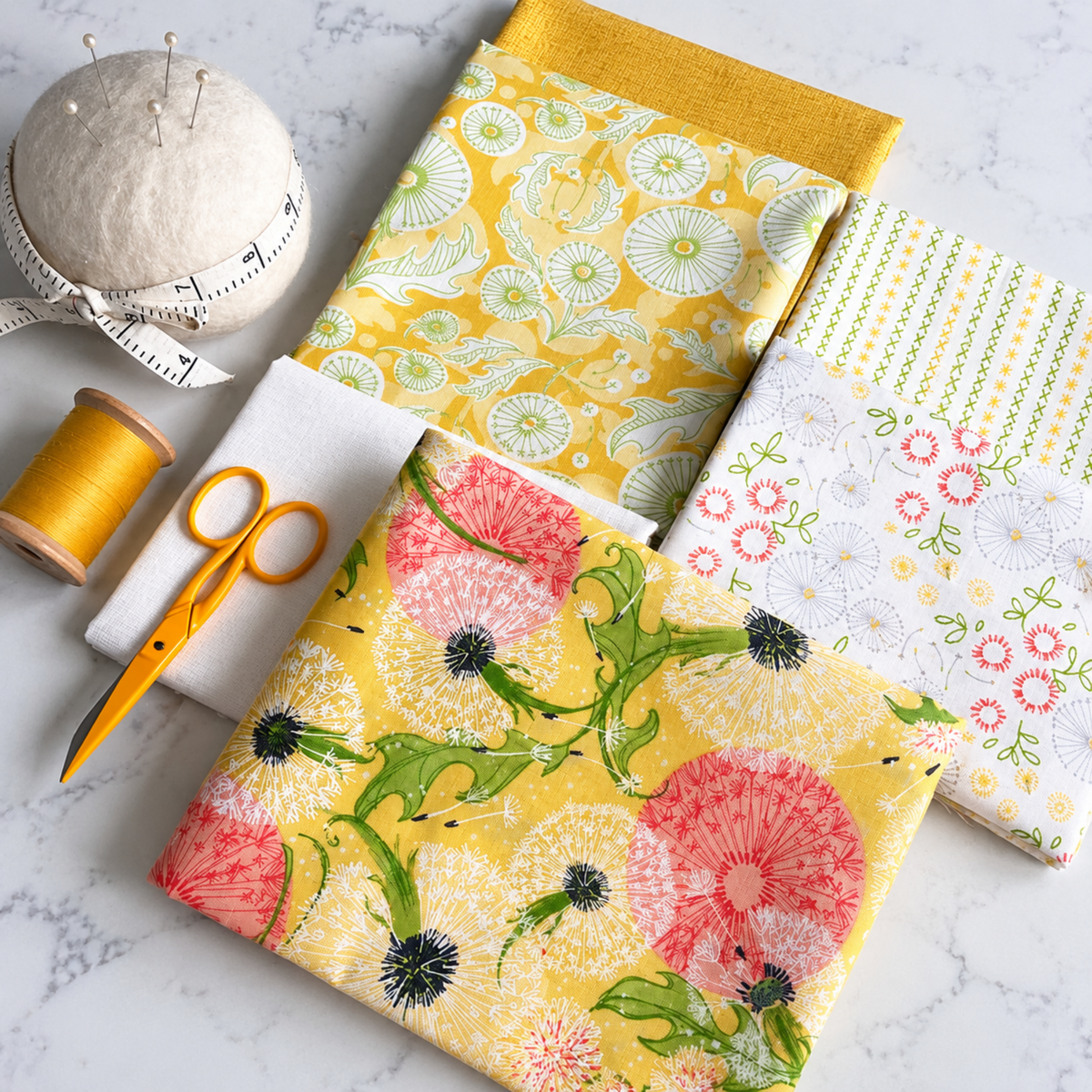 Folded yellow, cream, and floral dandelion-print fabrics beside thread, scissors, and a pincushion on a sewing table.