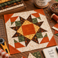 Interwoven Pumpkin Fabric by Jason Yenter for In The Beginning Fabrics as the center square and corner triangles in a Lone Star quilt block on a sewing table, with coordinating autumn prints, rotary cutter, and "Sew Quilt Be Happy" mug. Quilt blocks shown for inspiration. Coordinating fabrics and quilt patterns are available at IraRobi Quilts.