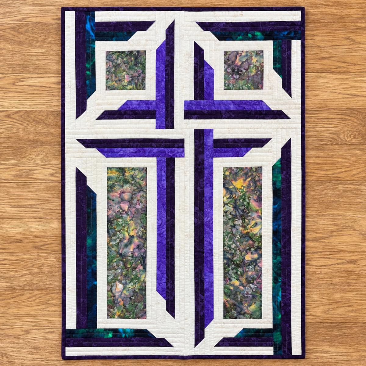 The Wondrous Cross handmade quilted wall décor with a white cross, purple and teal patchwork, and floral batik panels, laid flat on an oak floor.