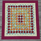Handmade scrappy 4-patch quilt in brick red, orange, yellow, and green tones, shown flat with a classic patchwork layout