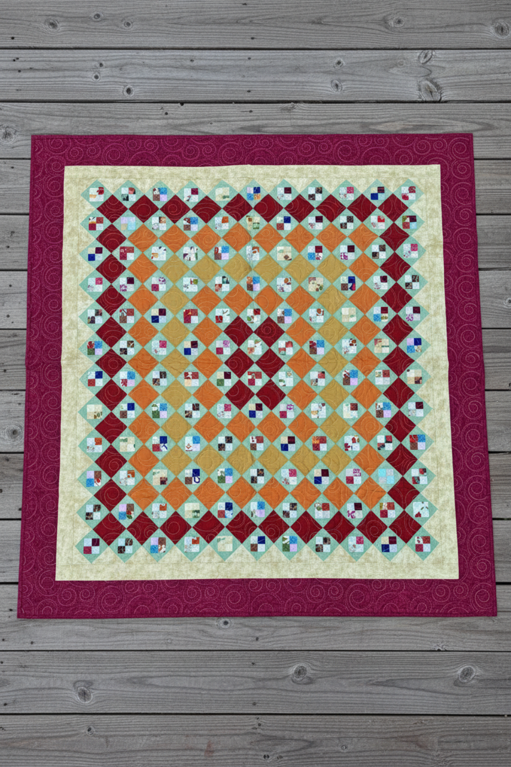Handmade scrappy 4-patch quilt in brick red, orange, yellow, and green tones, shown flat with a classic patchwork layout