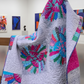 Colorful quilt with floral patterns displayed on a stand against a white wall with abstract art.