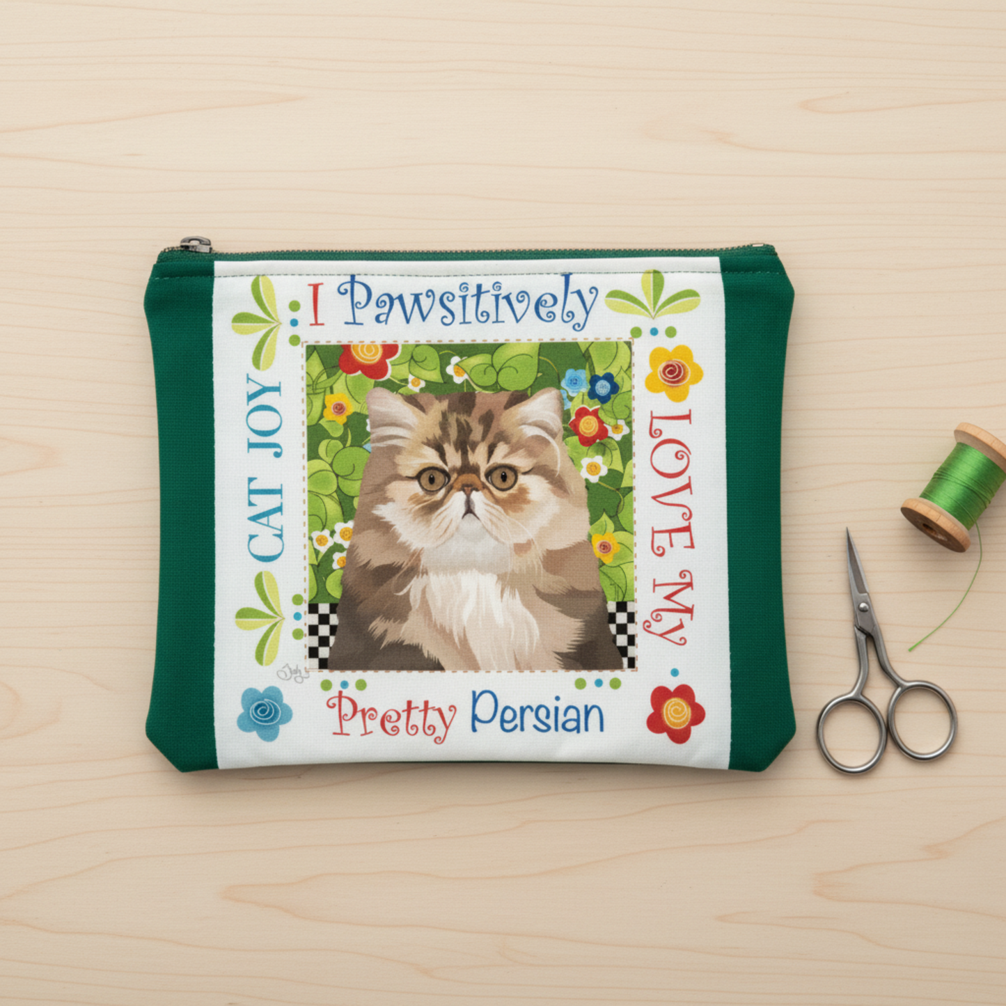 Pretty Persian Cat Joy fabric art panel by Jody Houghton Designs sewn into a green zippered pouch – project idea for 6" novelty panel, shown with scissors and green thread spool on wood surface