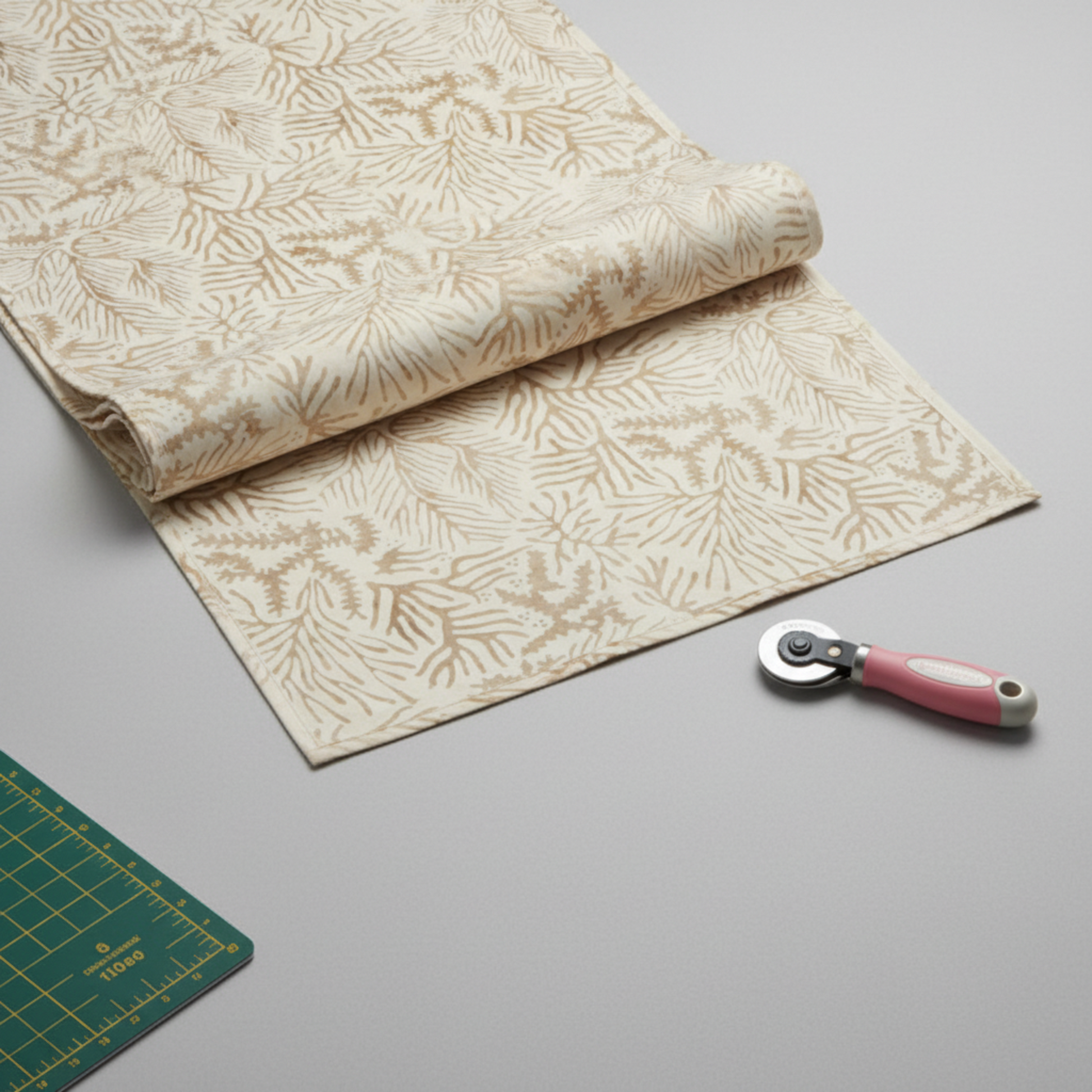 Expressions Batiks Breathe Ecru quilting cotton fabric by Riley Blake Designs – partially unrolled yard on light grey surface with pink rotary cutter and green cutting mat corner, warm cream and ecru tonal batik with pine needle and feathery frond motif in soft taupe and golden-beige