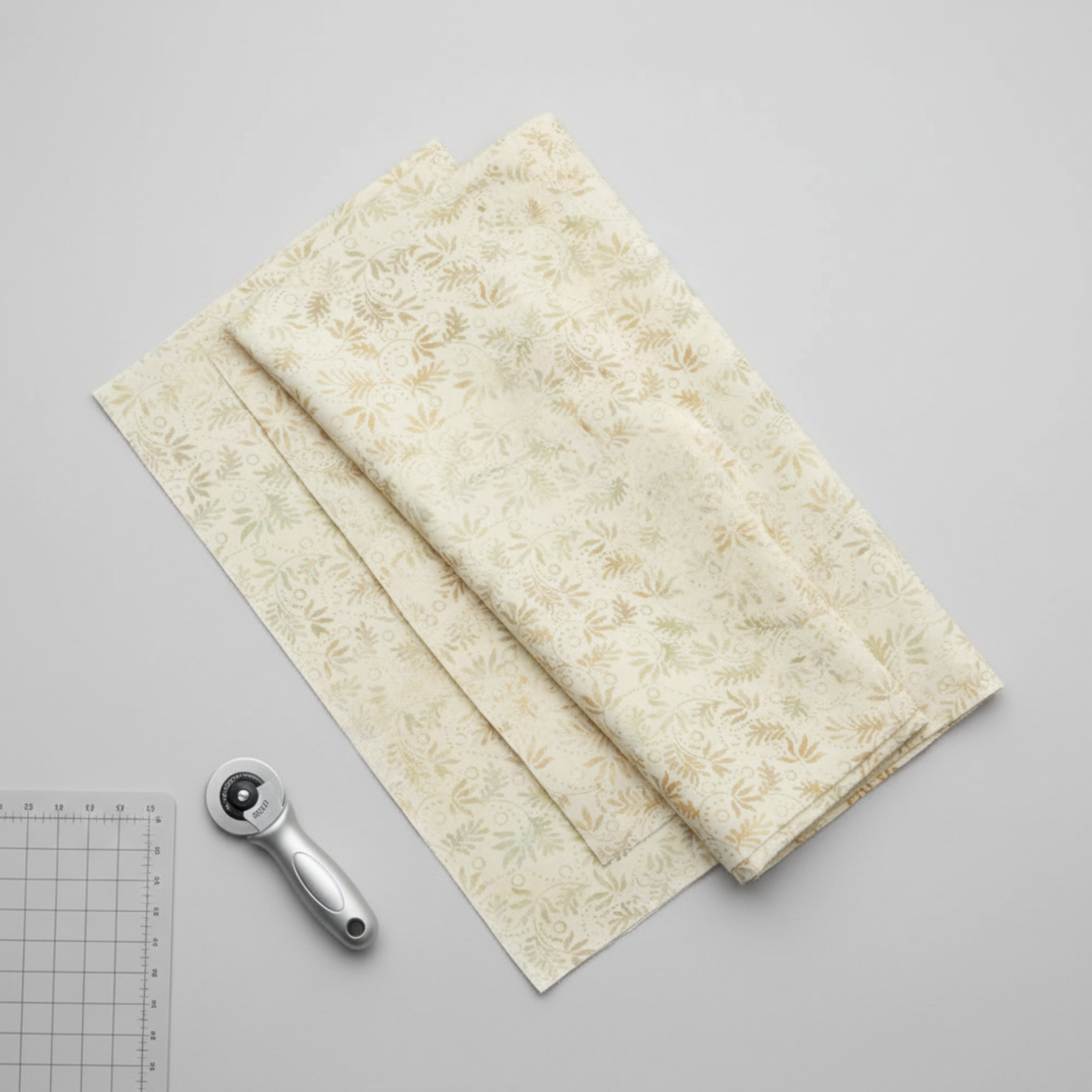 Expressions Batiks Breathe Laurel quilting cotton fabric by Riley Blake Designs – three accordion-folded fabric panels fanned on light grey surface with silver rotary cutter and small cutting mat corner, cream and ivory tonal batik with feathery fronds and leaf sprigs in soft sage green and warm taupe