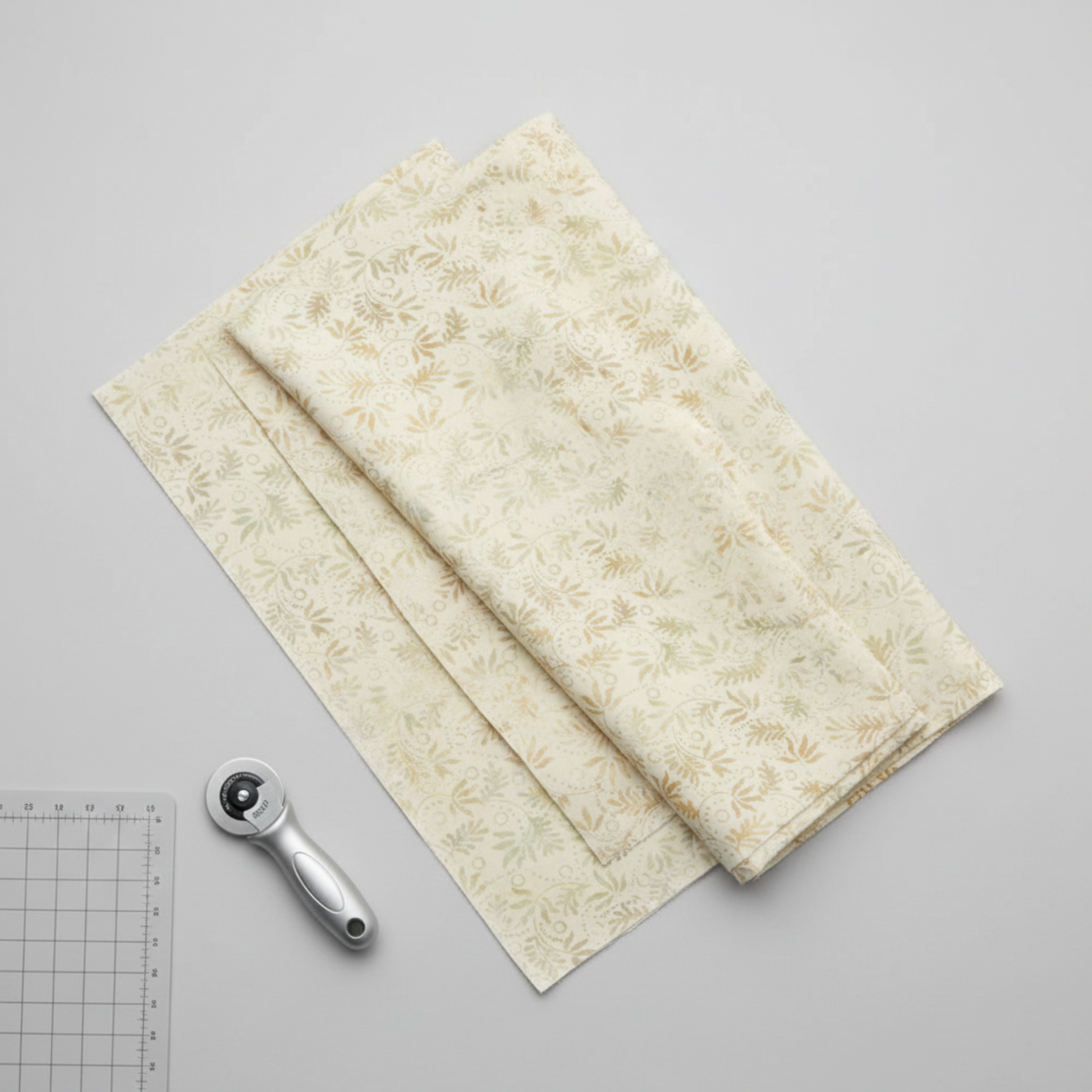 Expressions Batiks Breathe Laurel quilting cotton fabric by Riley Blake Designs – three accordion-folded fabric panels fanned on light grey surface with silver rotary cutter and small cutting mat corner, cream and ivory tonal batik with feathery fronds and leaf sprigs in soft sage green and warm taupe