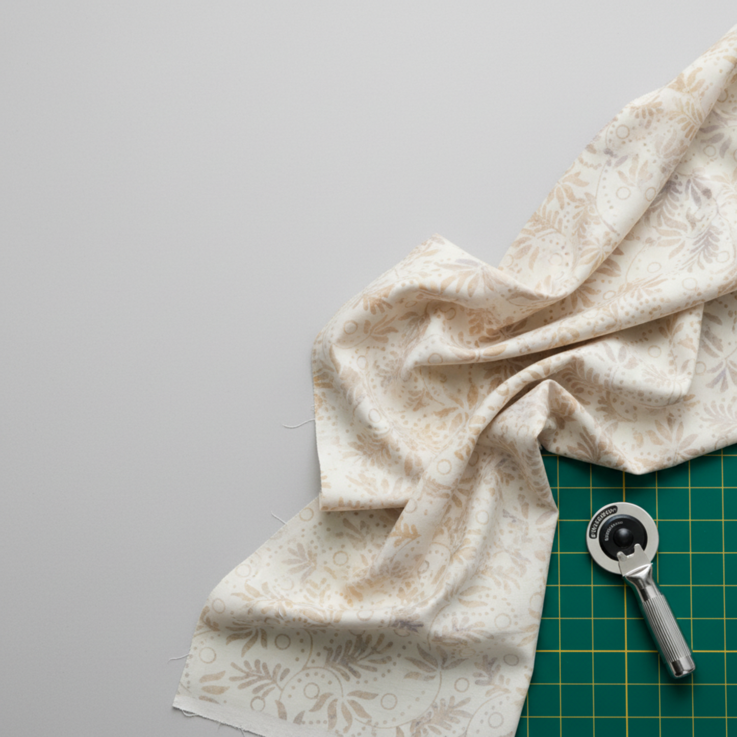 Expressions Batiks Breathe Flaxen by Riley Blake Designs – cream batik fabric loosely gathered on light grey surface with green cutting mat and rotary cutter, showing natural drape and soft hand in quilting workspace setting