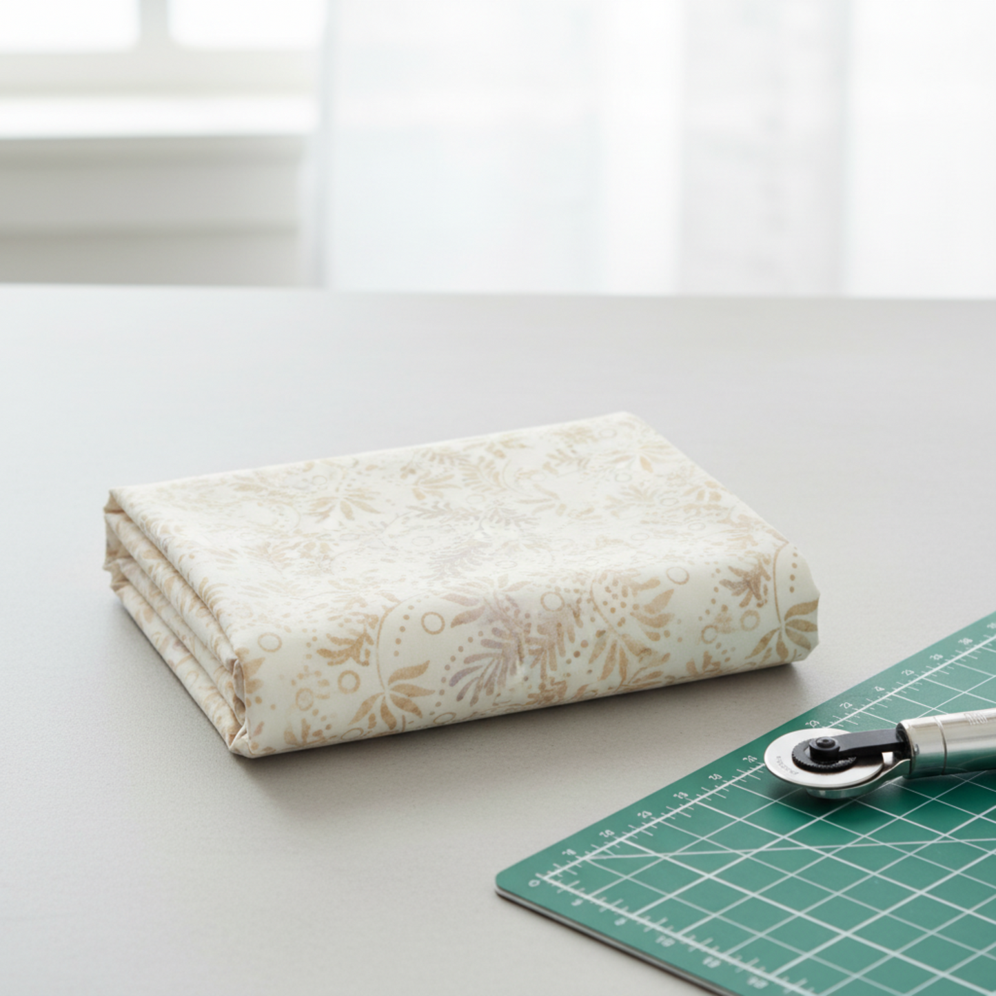 Expressions Batiks Breathe Flaxen by Riley Blake Designs – folded cream batik fabric on white surface beside green cutting mat and rotary cutter with soft natural window light, quilting workspace lifestyle shot