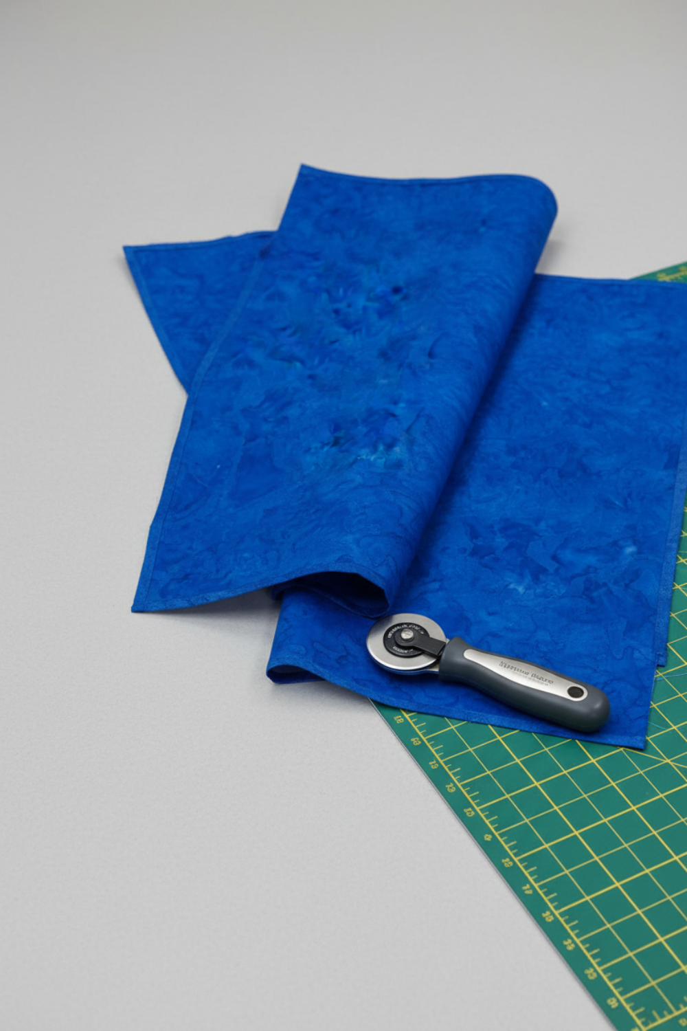 Expressions Batiks Hand-Dyed Cobalt quilting cotton with rotary cutter and edge of cutting mat on light grey background