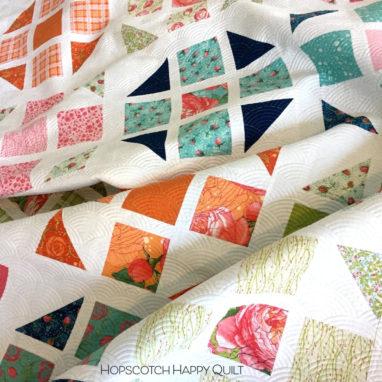 Hopscotch Happy quilt close-up flat lay showing colorful half square triangle blocks in orange, pink, teal, navy and floral prints on white background with scallop machine quilting by Robin Pickens