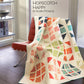 Hopscotch Happy quilt by Robin Pickens draped over red Barcelona chair by sunny window — colorful HST blocks in orange, pink, teal, green and navy on white background with teal binding