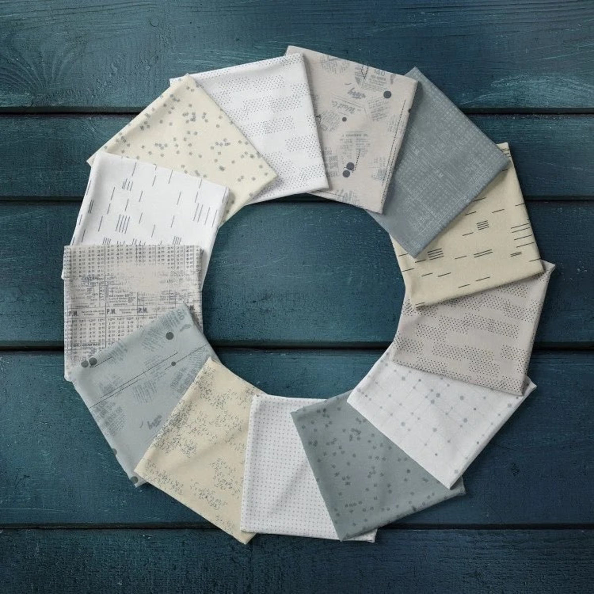 Folded Modern Background Even More Paper fabrics by Zen Chic for Moda Fabrics arranged in a circle, showing coordinating cream, eggshell, and steel-gray prints on a dark blue wooden surface.