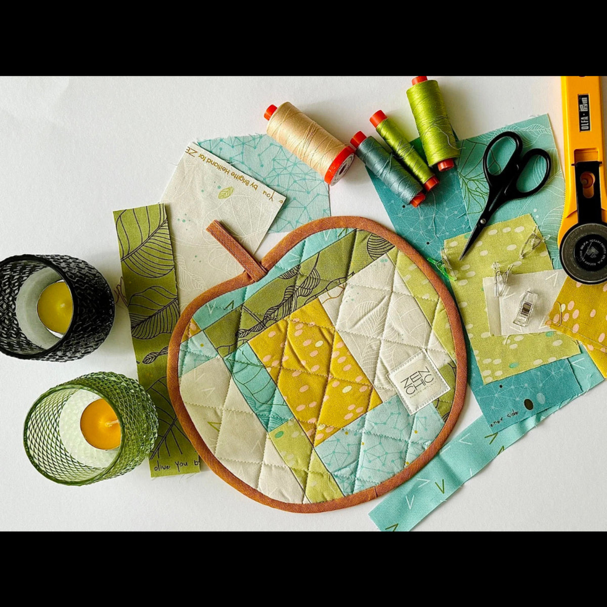 Overhead view of a round quilted project surrounded by Olive You fabric scraps in aqua, leaf green, cream, and yellow, with thread, scissors, and a rotary cutter.