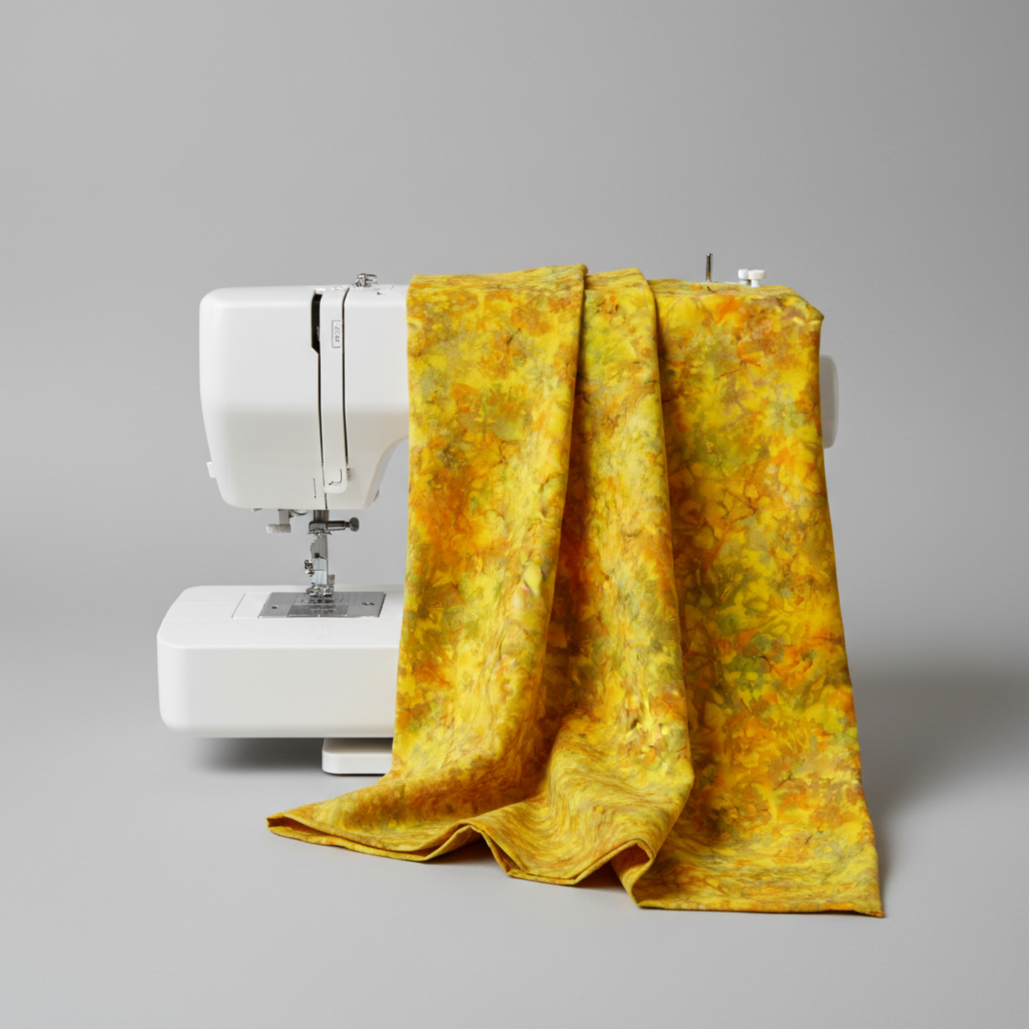 Expressions Batiks Hand-Dyed Yellow Orange Multi III quilting cotton fabric by Riley Blake Designs – vivid multicolor hand-dyed batik draped over white sewing machine showing natural drape and movement, golden yellow, orange, and olive green palette, light grey background