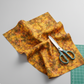 Expressions Batiks Hand-Dyed Orange Multi I quilting cotton fabric by Riley Blake Designs – loosely gathered on light grey surface with teal fabric scissors and green cutting mat corner, complex multicolor hand-dyed batik with golden amber-orange, olive green, and red-orange palette