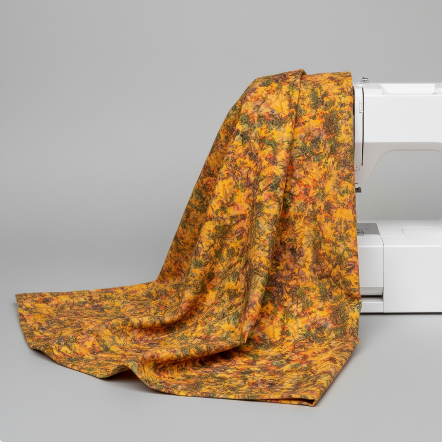 Expressions Batiks Hand-Dyed Orange Multi I quilting cotton fabric by Riley Blake Designs – draped over white sewing machine showing natural drape and movement, complex multicolor hand-dyed batik with golden amber-orange, olive green, slate blue-grey, and red-orange palette, light grey background