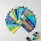 Tonga Batik Dreamer by Timeless Treasures – charm pack squares fanned on light grey background with Tonga Treats package, purple rotary cutter, and coordinating purple, teal, and yellow thread spools showing quilting workspace context