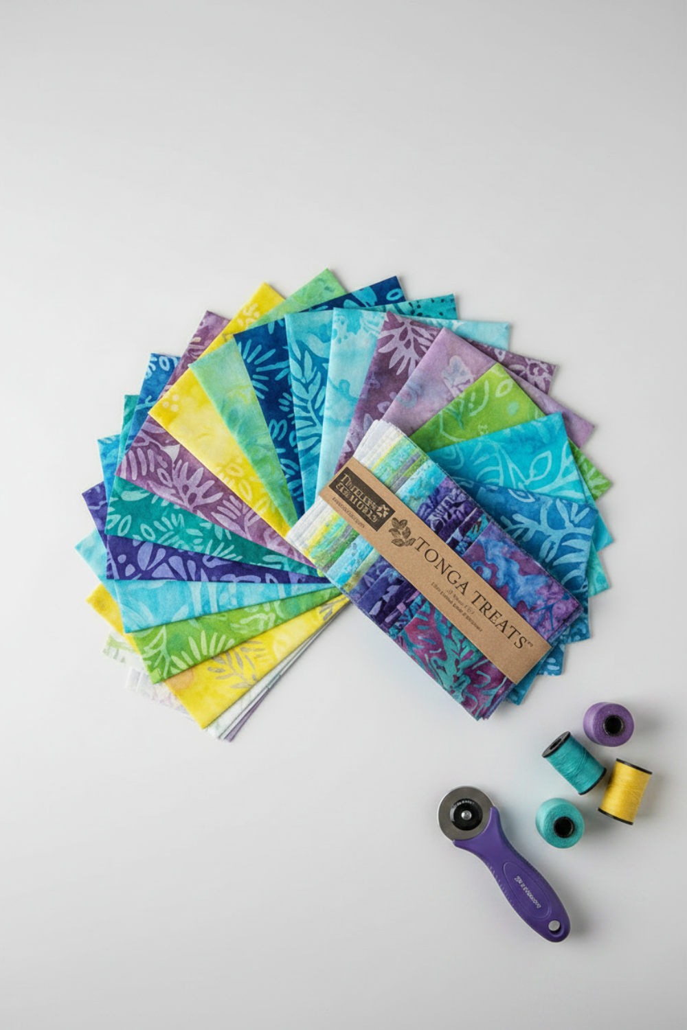 Tonga Batik Dreamer by Timeless Treasures – charm pack squares fanned on light grey background with Tonga Treats package, purple rotary cutter, and coordinating purple, teal, and yellow thread spools showing quilting workspace context