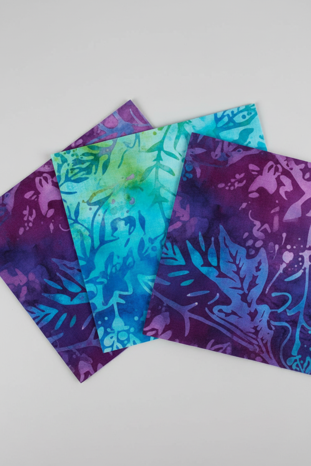 Tonga Batik Dreamer by Timeless Treasures – Pinterest close-up of three 5" charm squares fanned on light grey background showing vivid purple, teal, and blue-violet hand-dyed batik patterns with tropical leaf wax resist motifs