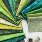 Artisan Batiks Prisma Dyes Rainforest by Robert Kaufman – Pinterest lifestyle shot of 40 pre-cut 2.5" strips fanned showing full green batik palette from cream and chartreuse through emerald, teal, and charcoal, with rotary cutter and green thread spool