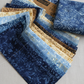 Tonga Batik Moonlit by Timeless Treasures – Pinterest lifestyle flat lay of 2.5" jelly roll strips fanned on natural linen surface with Tonga Treats package and folded deep navy daisy batik fat quarter, showing full palette of navy, soft blue, warm sand, and taupe hand-dyed batik