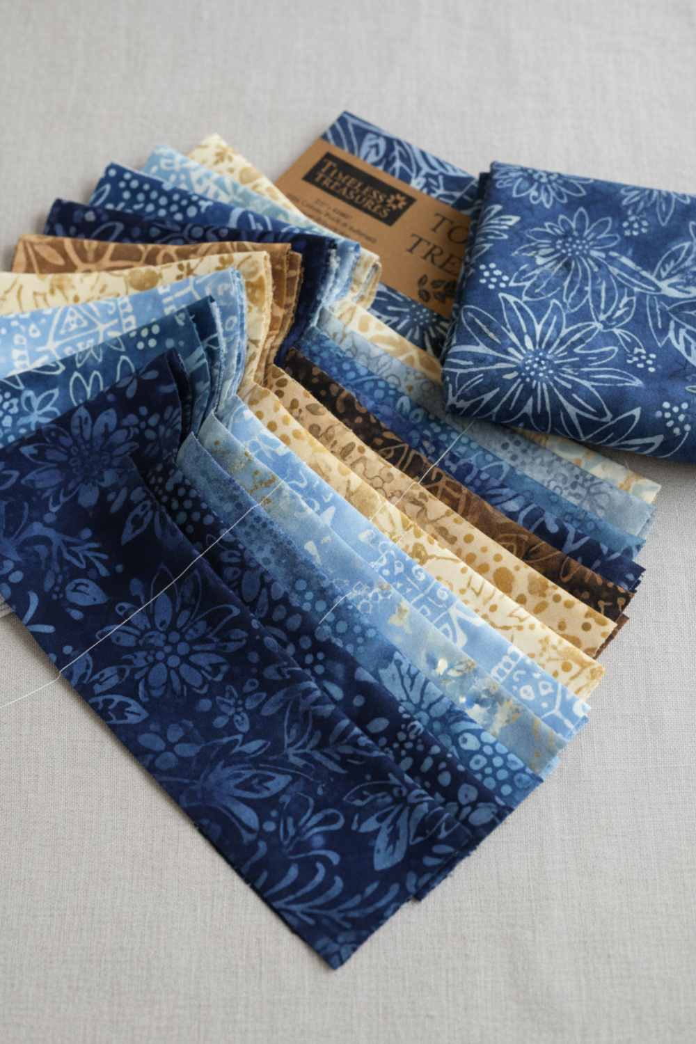 Tonga Batik Moonlit by Timeless Treasures – Pinterest lifestyle flat lay of 2.5" jelly roll strips fanned on natural linen surface with Tonga Treats package and folded deep navy daisy batik fat quarter, showing full palette of navy, soft blue, warm sand, and taupe hand-dyed batik
