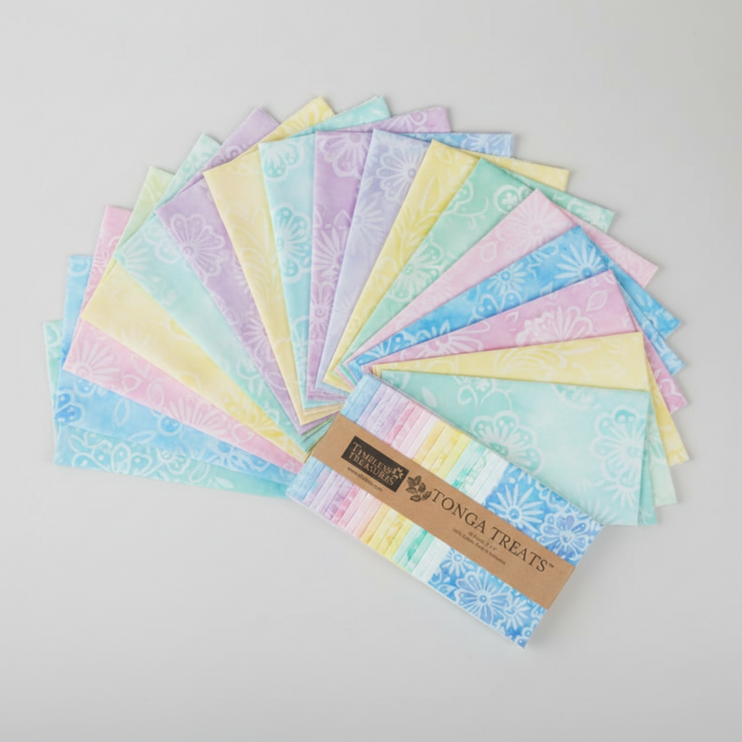 Tonga Batik Pixie by Timeless Treasures – 42 pre-cut 5" charm squares fanned in wide arc on light grey background with Tonga Treats package, showing full pastel rainbow palette of mint, aqua, sky blue, lavender, blush pink, and soft yellow hand-dyed batik with floral motifs