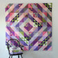 Faceted quilt pattern by Robin Pickens — purple, pink and green half square triangle quilt displayed on white wall with dark Windsor chair and folded quilts
