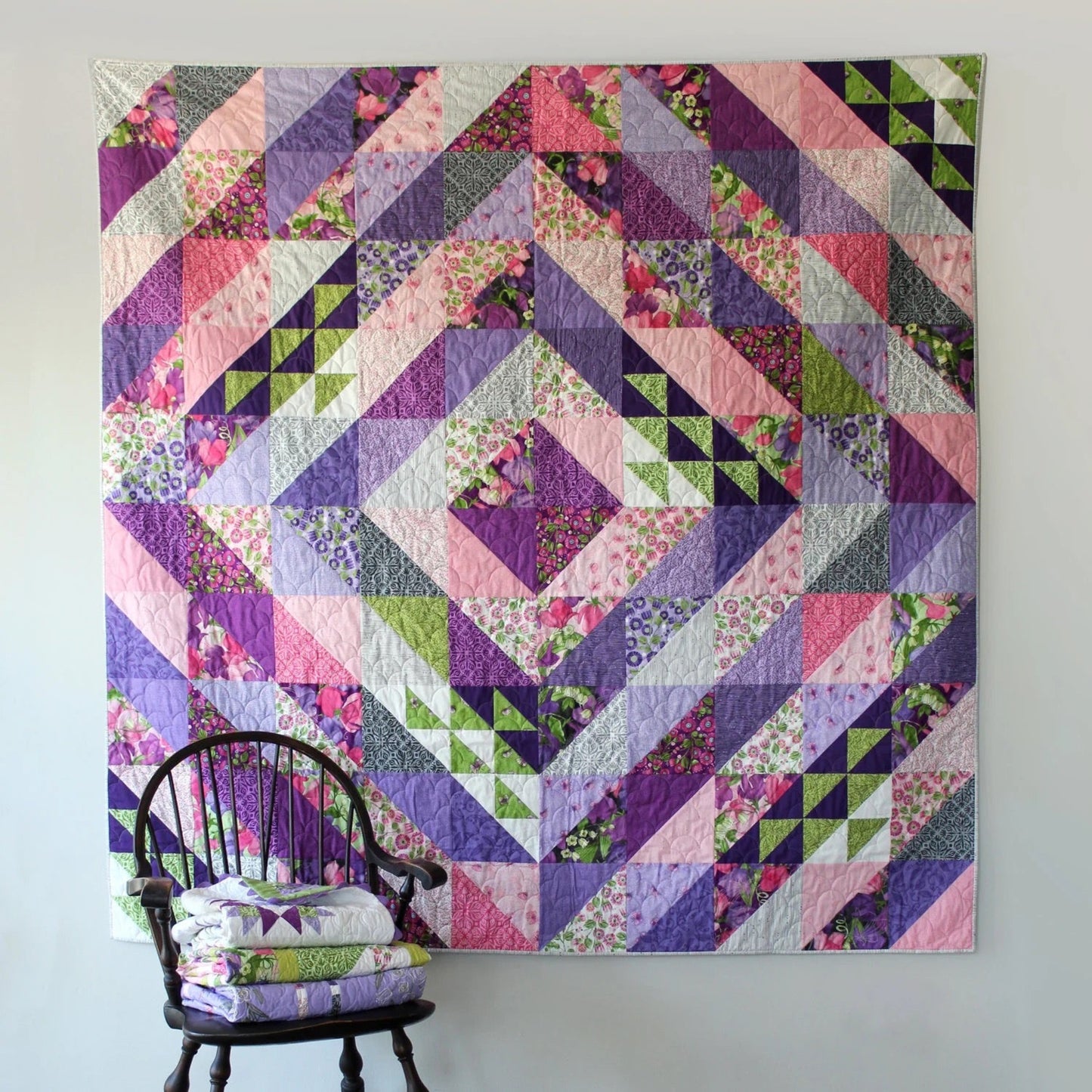 Faceted quilt pattern by Robin Pickens — purple, pink and green half square triangle quilt displayed on white wall with dark Windsor chair and folded quilts