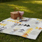 Writing on the Wall quilting cotton collection by FreeSpirit Fabrics -- Spotted Geese quilt spread on summer grass as a picnic blanket, featuring flying geese blocks in black word collage, white word collage, and golden yellow abstract scratch print fabrics on a white background, styled with a wicker picnic basket, ceramic mugs, and stacked plates