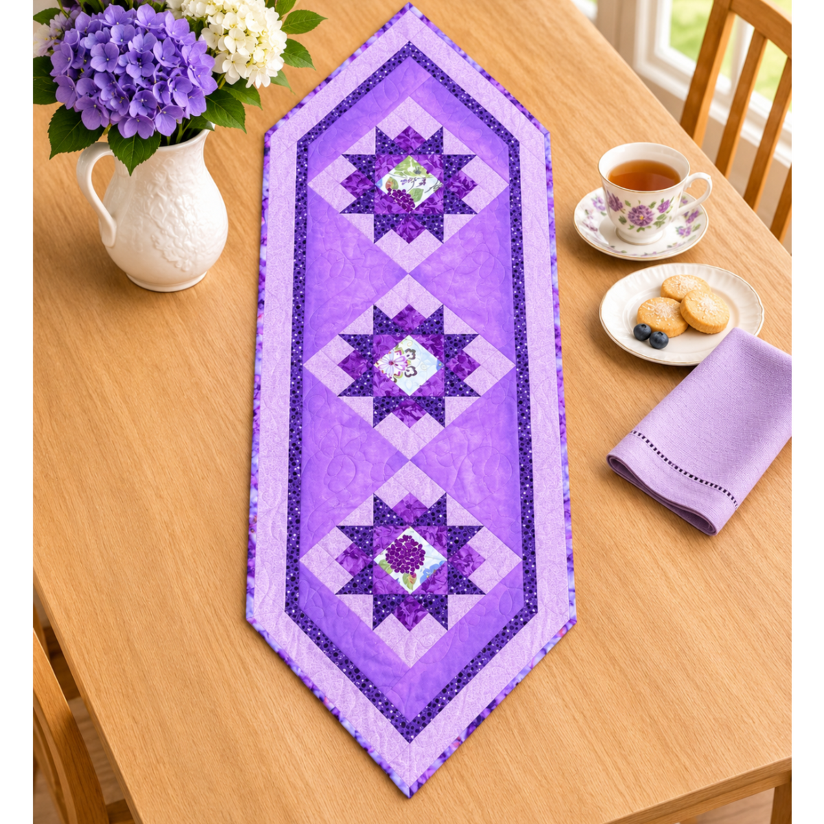 Purple Twilight quilted table runner on a wood dining table beside purple and white hydrangeas, a floral teacup, cookies and a lavender napkin.