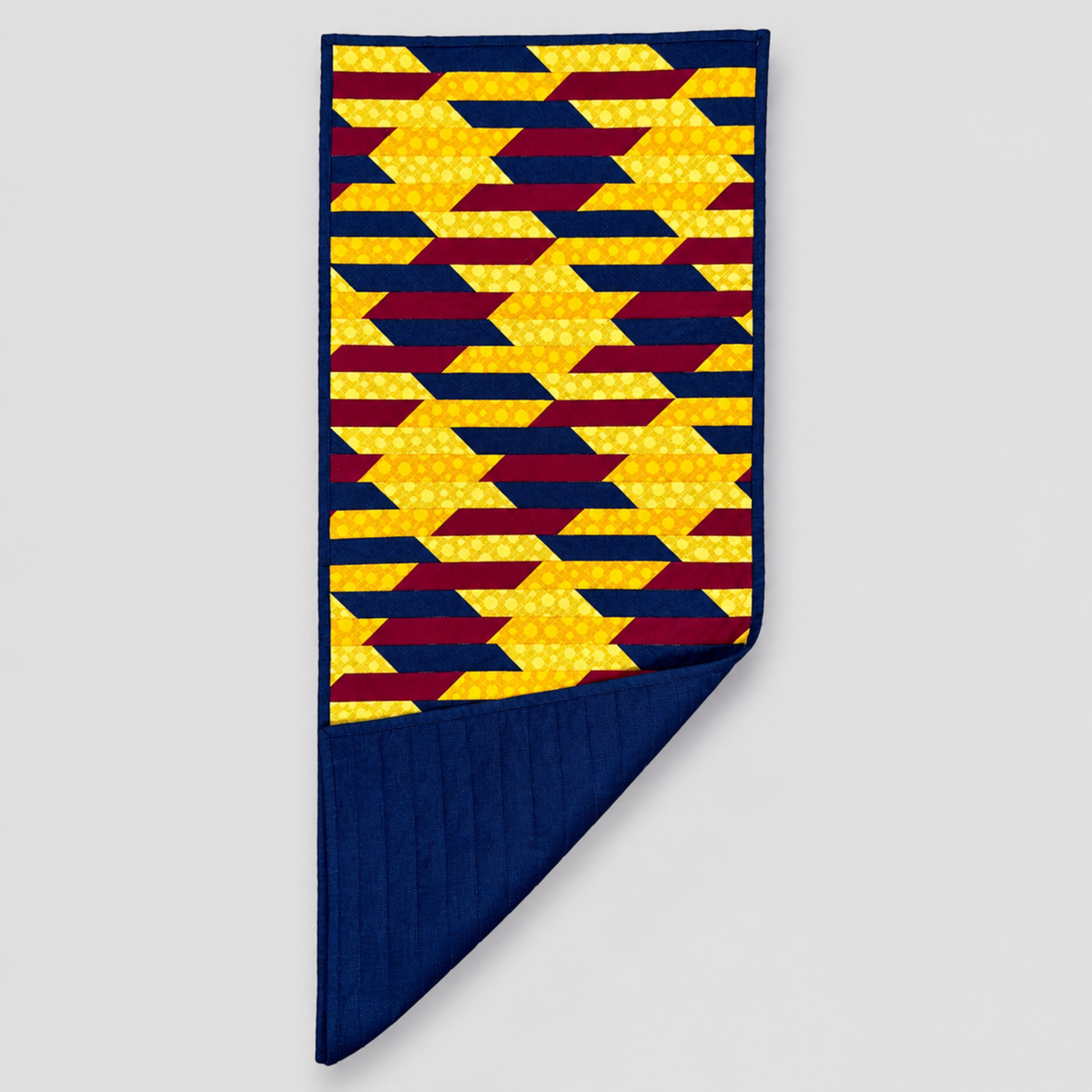 Golden Weave quilted table runner with one corner folded back to reveal navy backing and quilting beneath golden yellow, navy and burgundy patchwork.